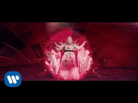 Music video scene featuring a singer in a floral gown surrounded by dancers against a backdrop of red and geometric patterns.