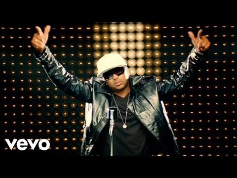Music video scene features a singer in a black jacket, baseball cap, and sunglasses, singing into a microphone against a lit backdrop.