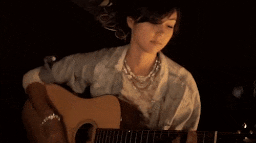 Young woman with curly brown hair sits indoors playing an acoustic guitar; she wears a light blue denim jacket and layered pearl necklaces.
