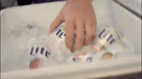 Close-up of a hand reaching into a white cooler filled with ice to grab a can of beer.