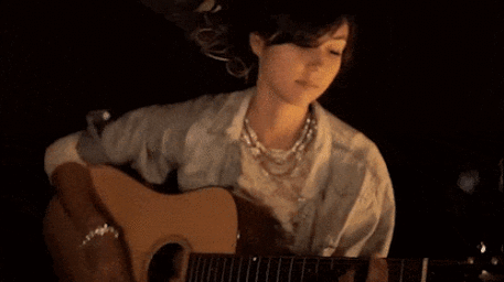 A woman playing an acoustic guitar wearing a light denim shirt and layered beaded necklace against a dark background.