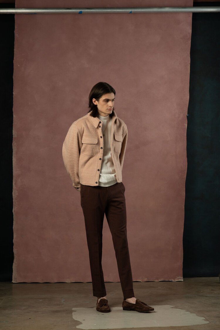 A young adult male poses in a pink jacket and brown trousers against a muted background, showcasing a blend of casual and formal fashion.