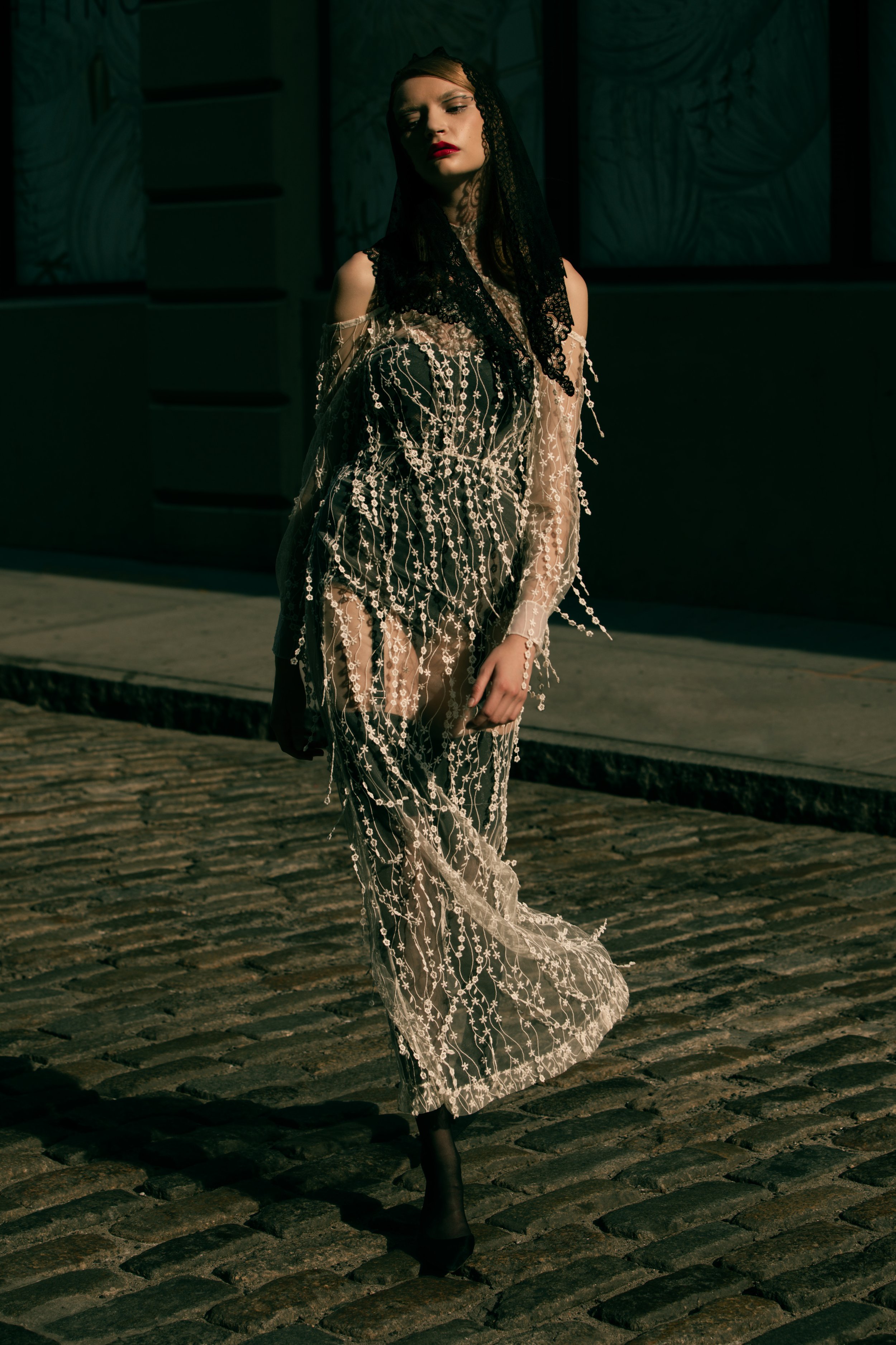 A lone woman in a pearl-beaded sheer gown and dark veil walks on a cobblestone street in daytime, shadowed by a tall building.