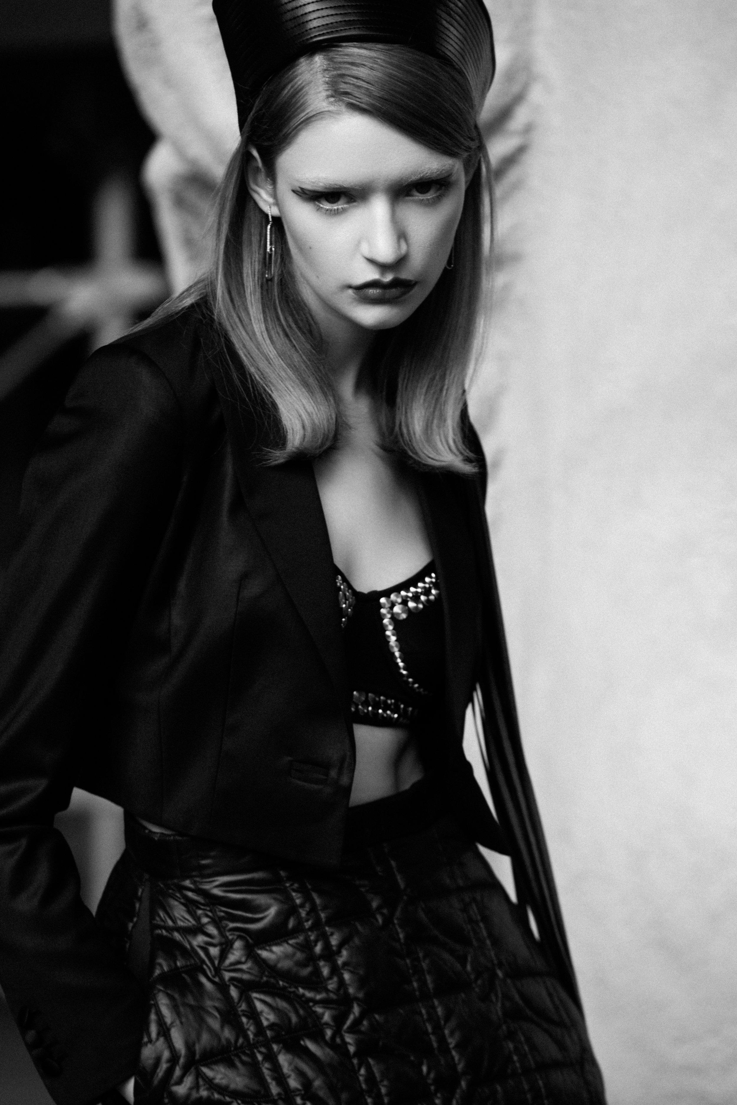 Black-and-white fashion portrait of a stylish young woman in a cropped blazer, beaded bra top, and quilted skirt, posing in a studio.