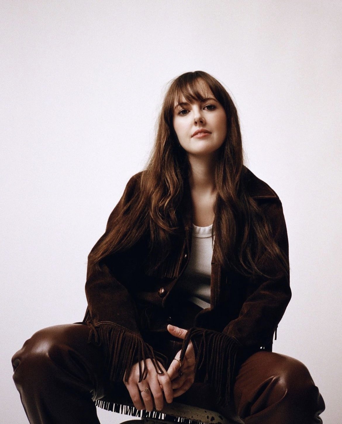 A woman with long brown hair poses in a brown suede jacket and leather pants, seated against a white background in a medium shot.