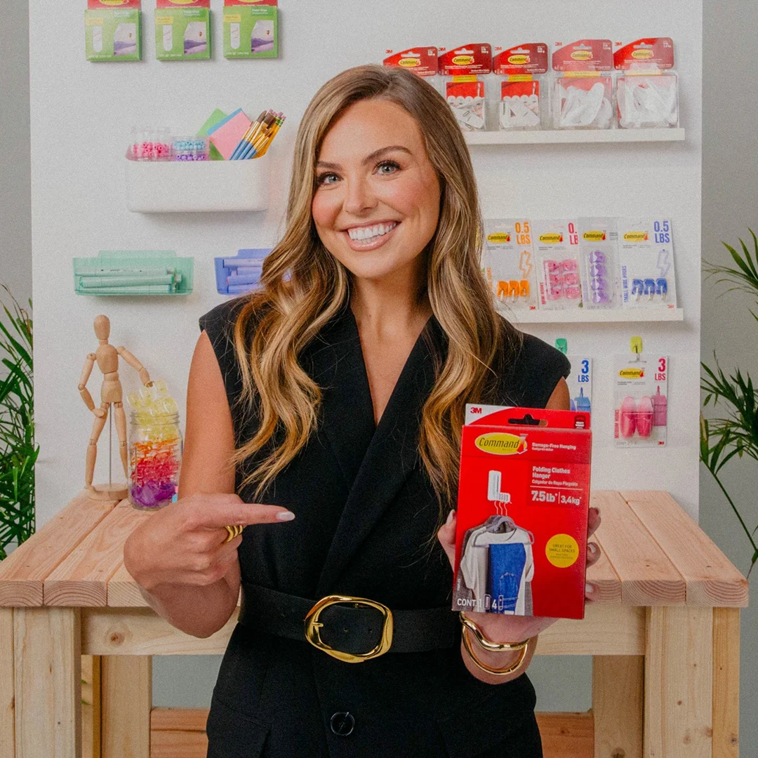 Adult woman smiles and points to a red package labeled 'Command Folding Hanger' in an indoor setting with shelves and objects.