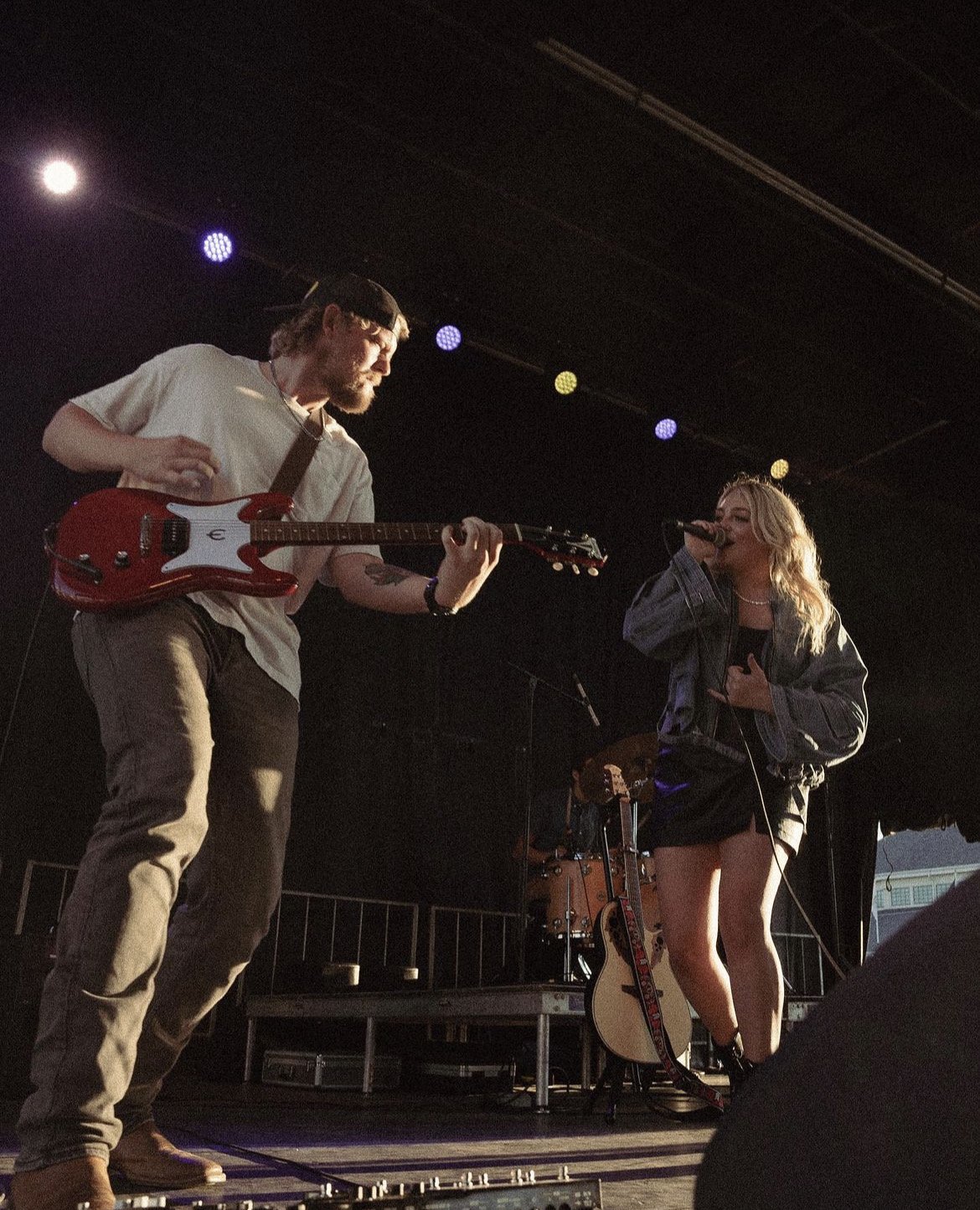 Live music performance featuring a male guitarist and female singer on stage, illuminated by stage lights against a dark backdrop.