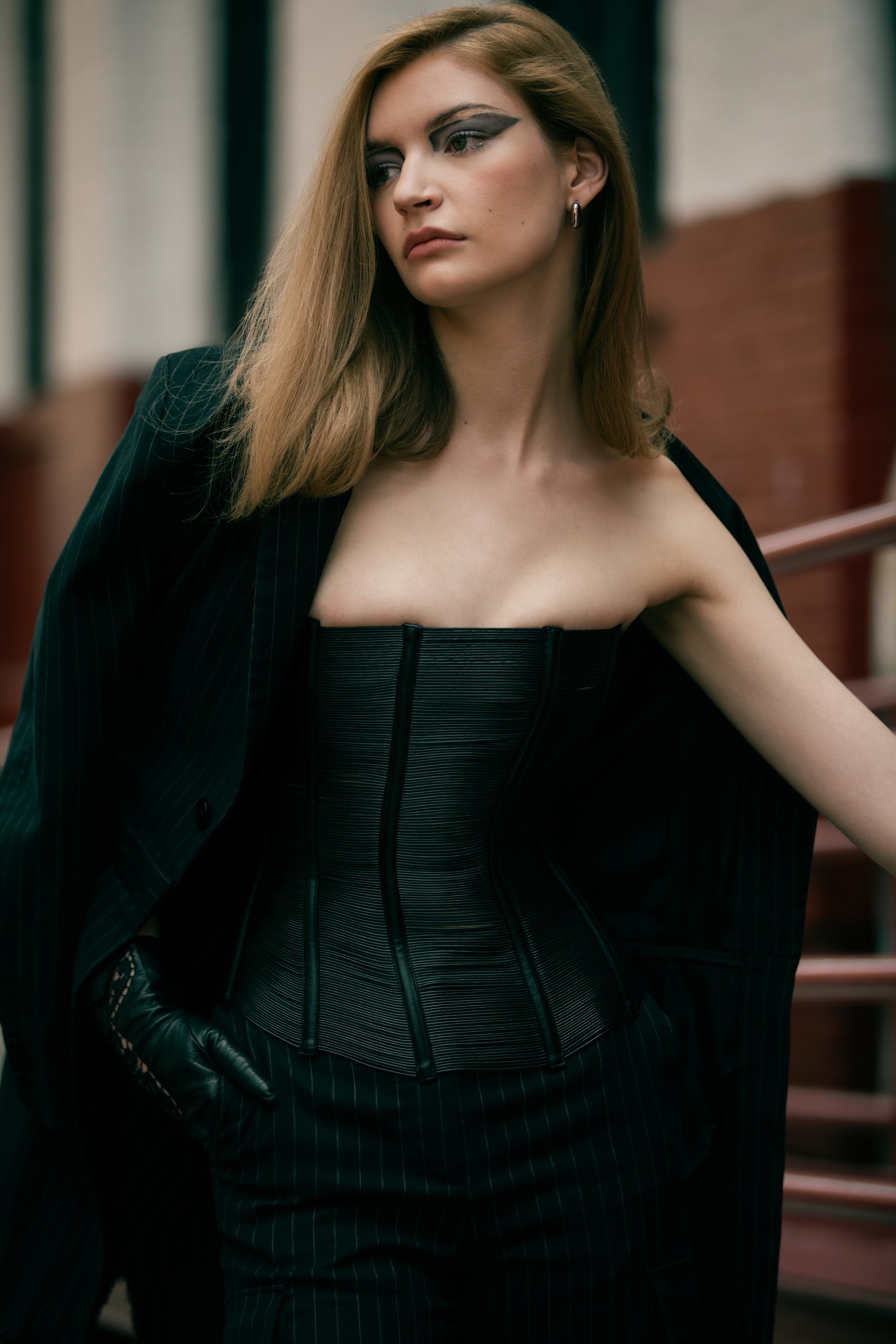 A young adult woman posing outdoors in a dark corset, blazer, and trousers ensemble with dramatic eye makeup and neutral hair.
