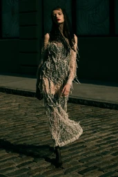 Model in a sheer fringed dress and black lace head covering walking on a cobblestone street.