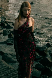 Model in dark red-spotted gown and long black gloves standing by rocky shore