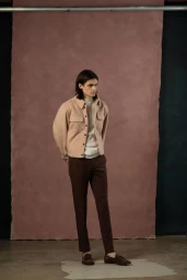 Full-length portrait of a male model wearing a light pink button-up jacket, white knit sweater, and brown trousers.