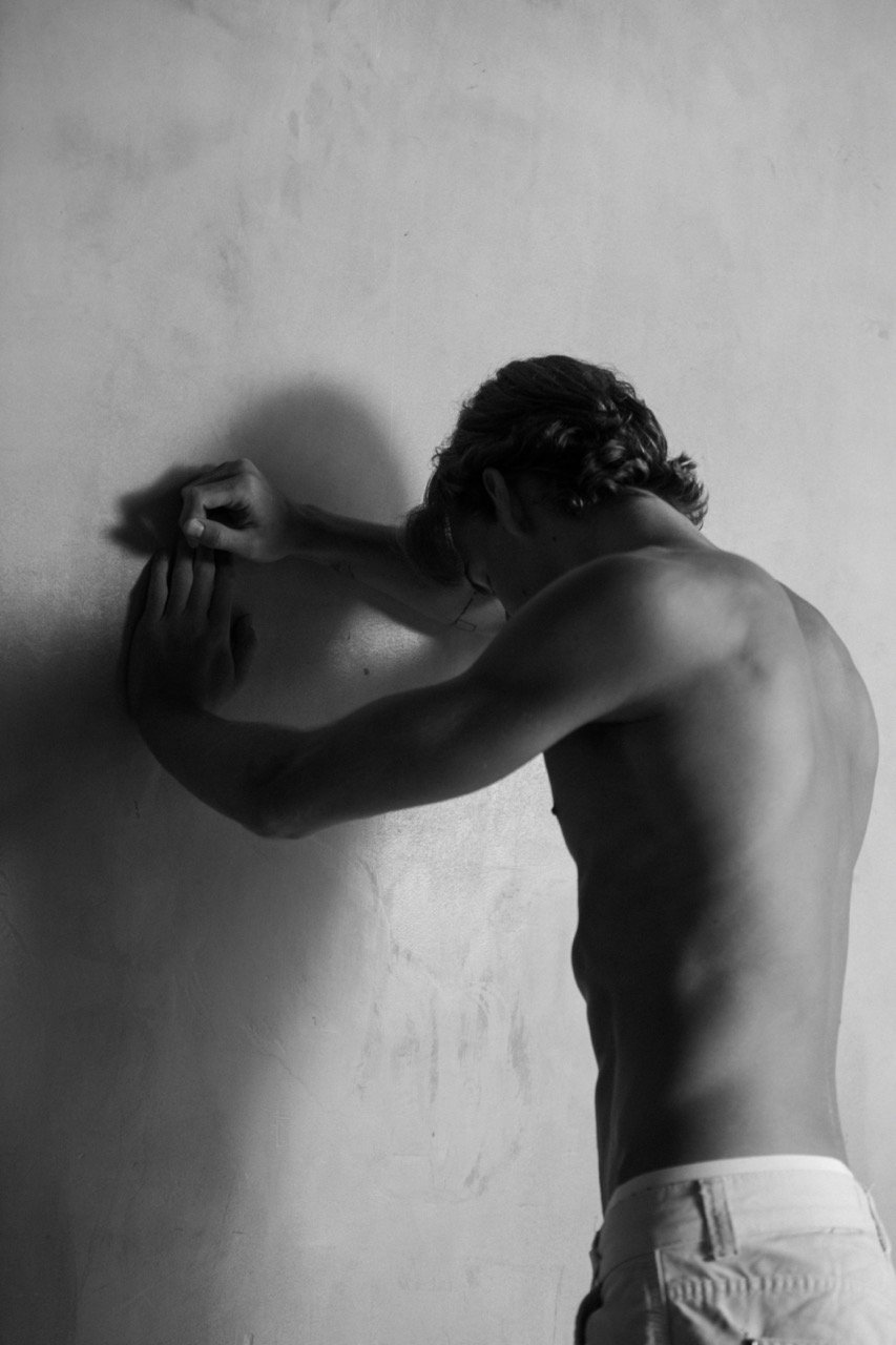 Black and white portrait of a shirtless man leaning against a wall, showcasing his physique in a minimalist setting with soft lighting.