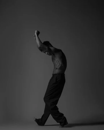 Shirtless man wearing dark trousers posing with arms raised against a grey studio backdrop