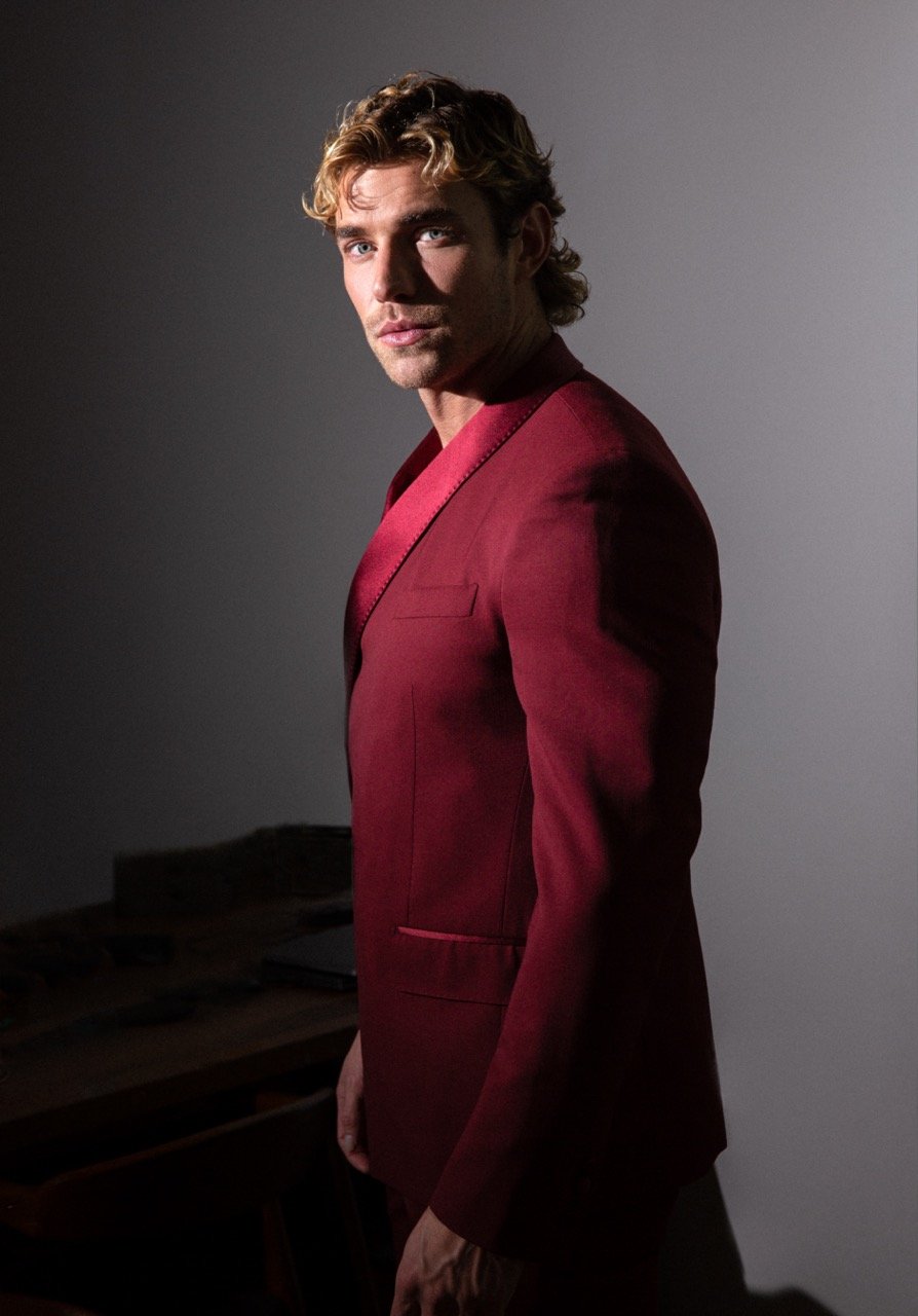 Portrait of a handsome man with wavy blond hair wearing a red suit, set against a studio backdrop with low-key lighting.