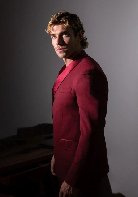 Portrait of a handsome man with wavy blond hair wearing a red suit, set against a studio backdrop with low-key lighting.