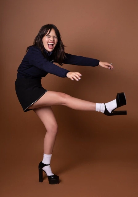 Woman with dark hair poses in a navy long sleeve top, black mini skirt, white socks, and black platform heels against a brown backdrop.