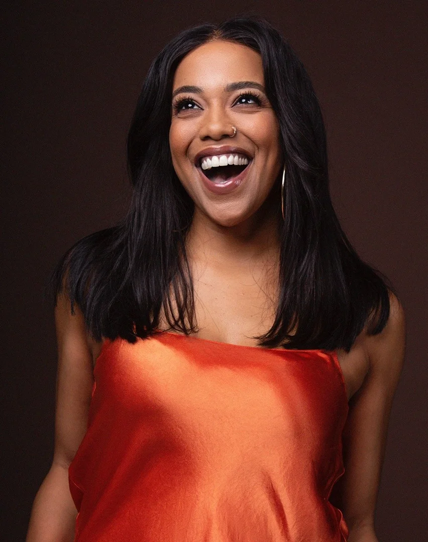 A close-up studio portrait of a smiling adult female with dark hair and an orange dress against a warm brown backdrop, looking upward.