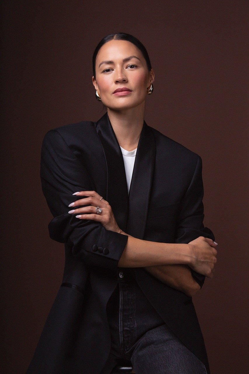 Portrait of an adult woman with her arms crossed, wearing a black jacket and jeans, posing against a brown background in a studio setting.