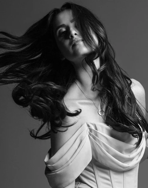 Black and white portrait of a young woman with long flowing hair wearing an off-the-shoulder top in a grey studio setting.