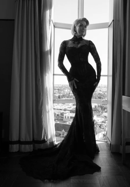 Black and white portrait of an adult woman in a lace gown, standing in front of an open window with a cityscape view.