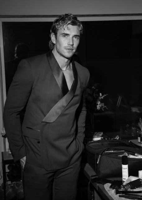 Black and white portrait of a man in a suit, looking at the camera, with a dimly lit background and various objects.
