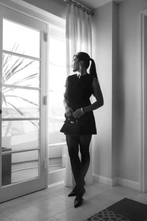 Woman in dark sleeveless dress standing by a balcony door holding a small handbag