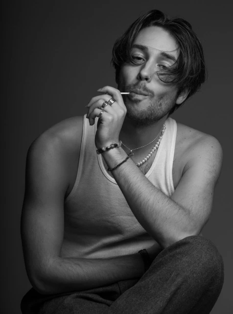 Black and white portrait of a young adult male in a tank top with dark hair, seated and holding a lollipop, against a grey backdrop.
