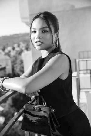 Monochrome portrait of a young woman leaning on a balcony railing, holding a dark handbag.