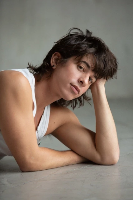 Portrait of a young adult with dark hair in a white tank top, lying down in a light gray environment.