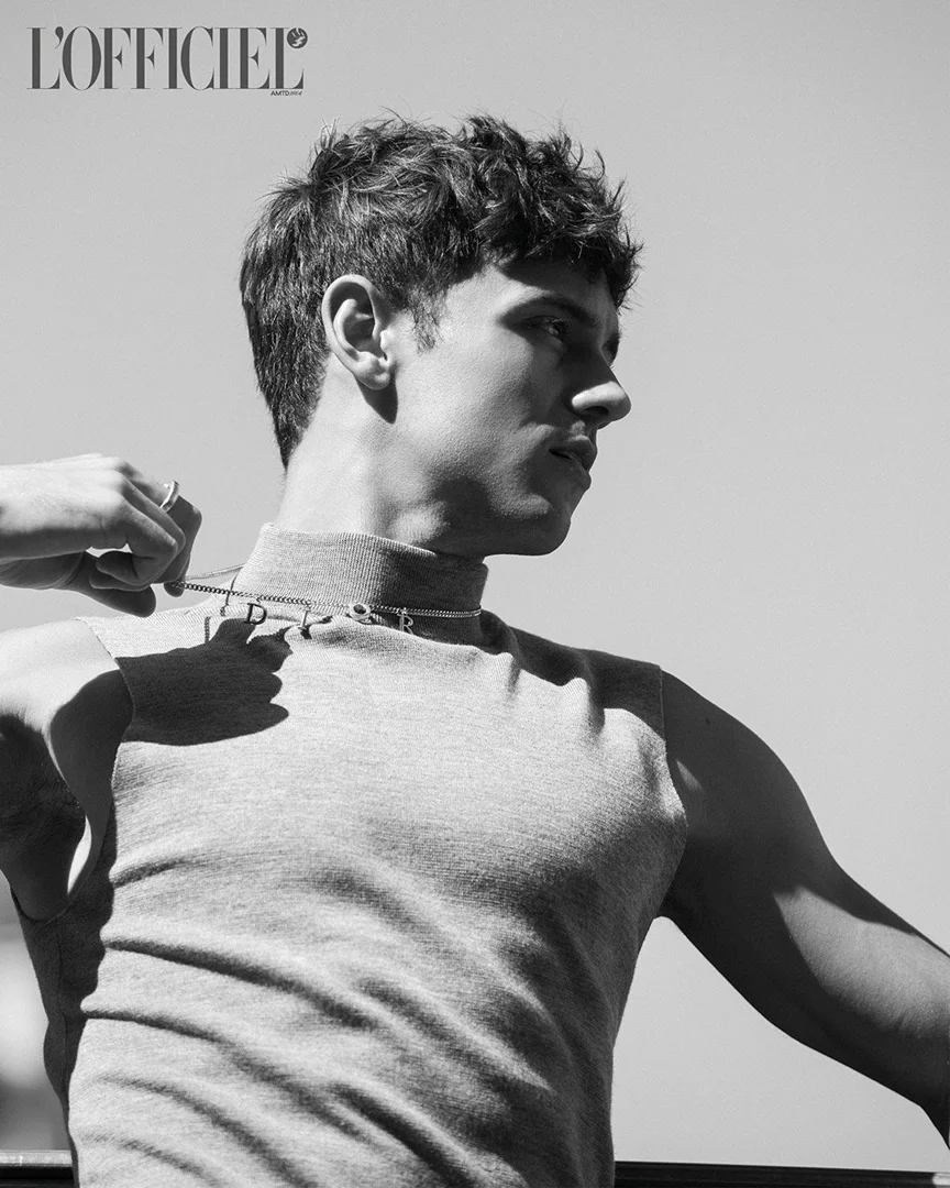Black and white portrait of a young adult male in a sleeveless top adjusting a necklace, with a clear sky background and magazine logo.