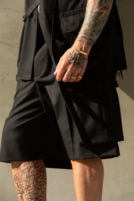 Man in avant-garde fashion poses with arm tattoos, wearing a black layered outfit against a neutral background, showcasing unique style.