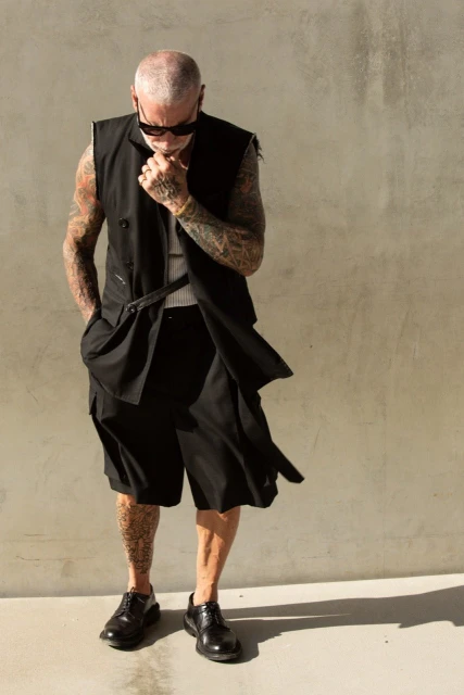 Stylish man with tattoos, sunglasses, vest, and shorts poses outdoors against a textured wall in full sunlight.