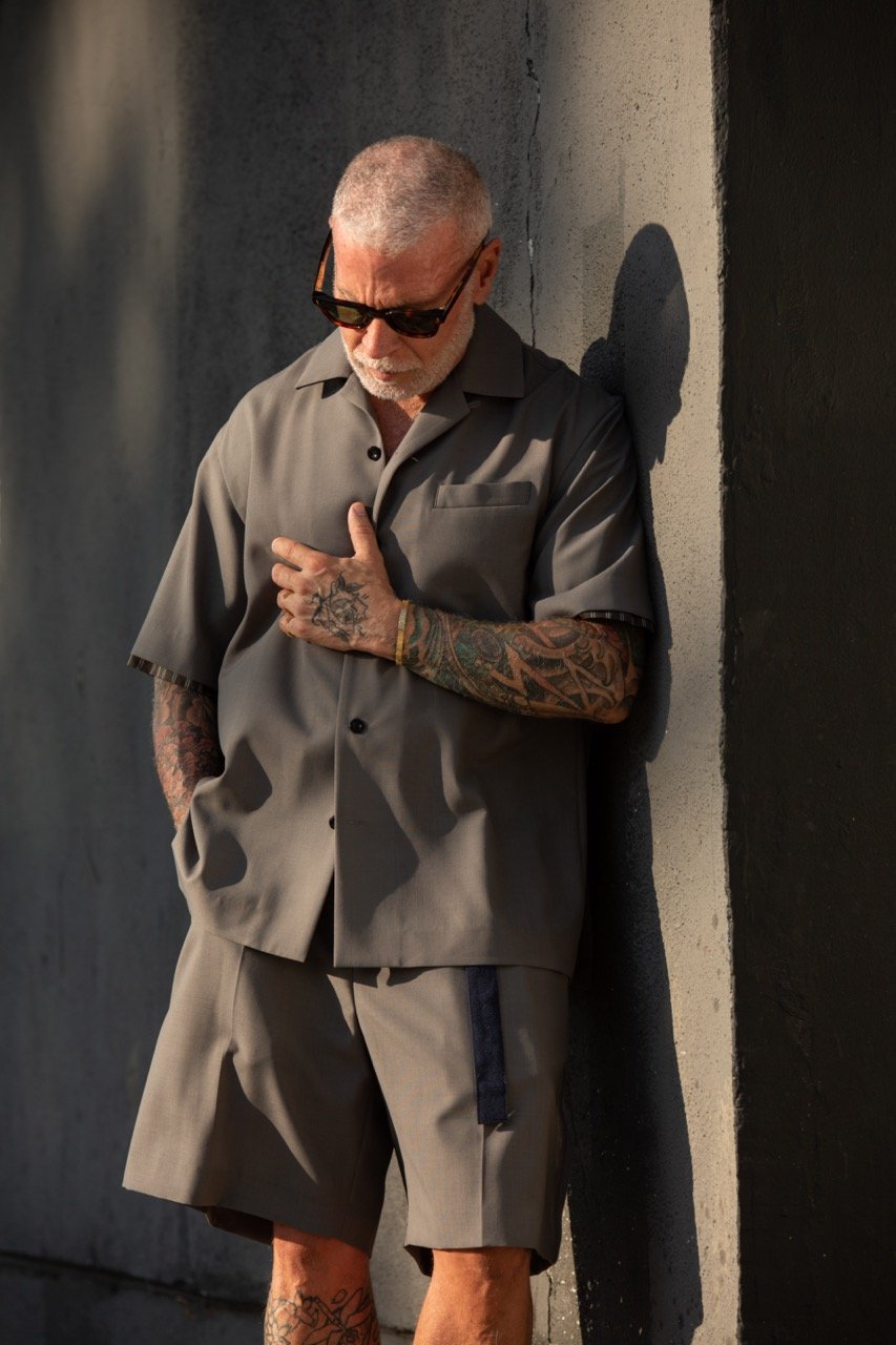 Adult man with tattoos poses against a textured wall in a matching gray shorts and button-down shirt, wearing sunglasses, looking down.