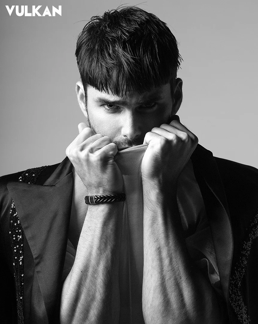 Black and white portrait of a stylish man with dark hair, wearing a blazer and holding a white garment near his mouth in a studio.