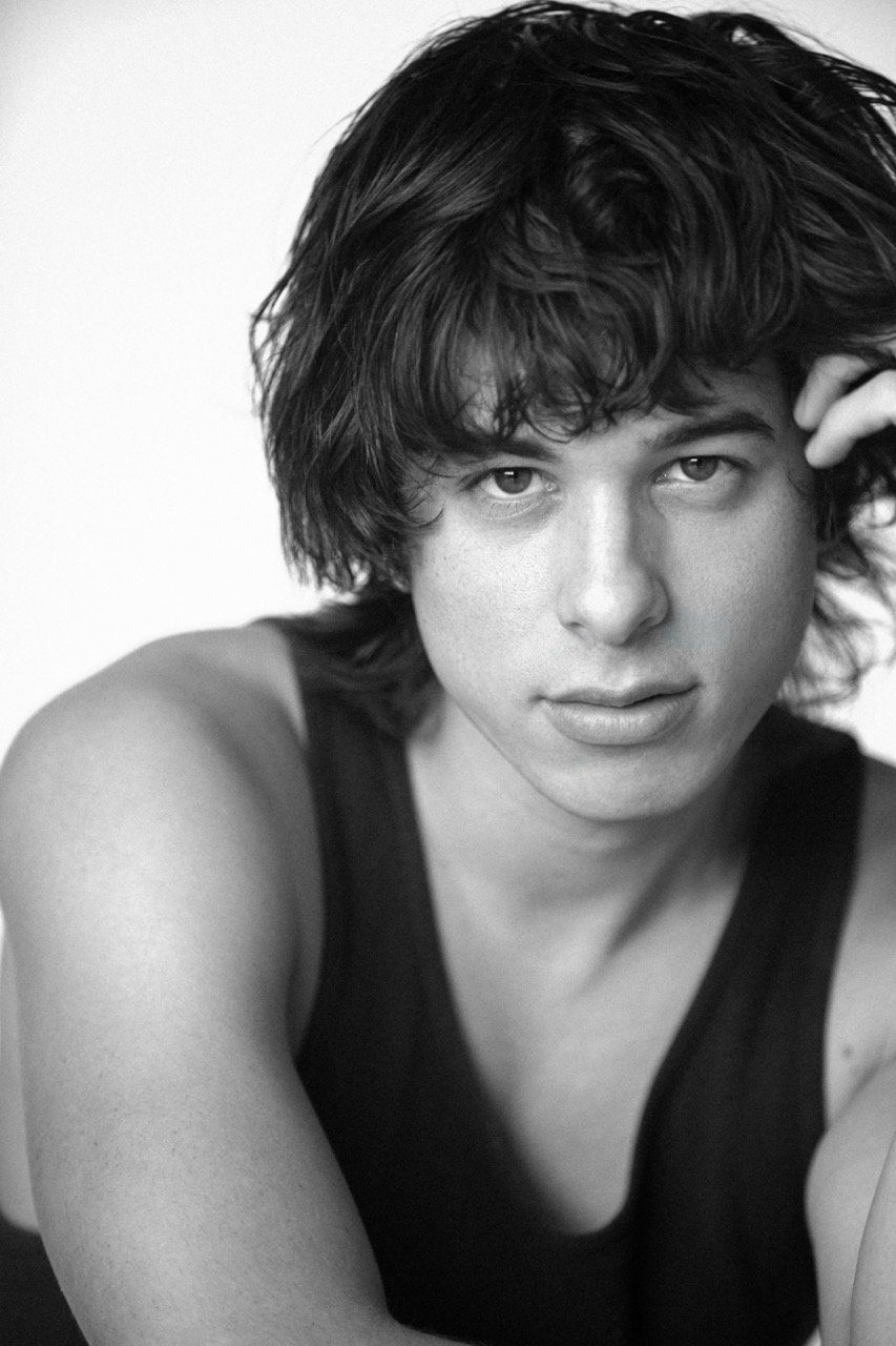 Monochrome portrait of a young adult man with dark hair, looking directly at the camera in a sleeveless top, against a white backdrop.