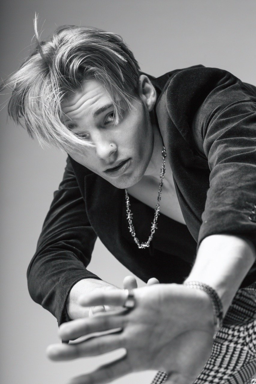 Monochrome portrait of a young man wearing a dark blazer and patterned trousers reaching toward the camera