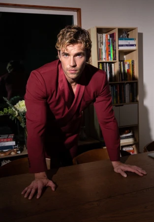 Man with curly blonde hair wearing a maroon jacket leaning over a wooden table inside a room with a bookcase.