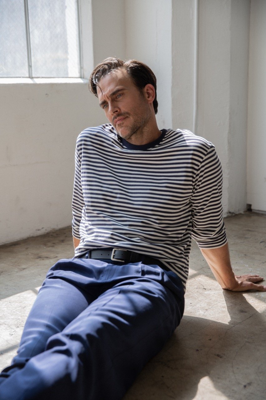 Man in a striped long-sleeve shirt and blue trousers sitting on a concrete floor near a window.