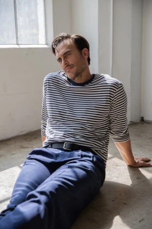 Man in a striped long-sleeve shirt and blue trousers sitting on a concrete floor near a window.