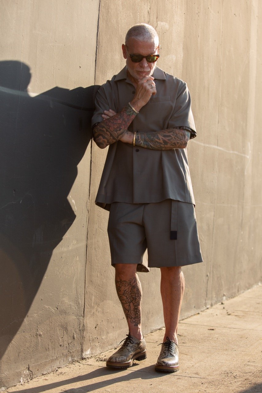 Mature man in a gray short set and sunglasses poses against a textured wall, showcasing his tattoos and stylish look in an outdoor setting.