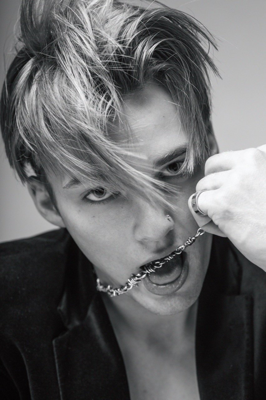 Black and white portrait of a young adult male with bleached hair holding a metal chain in his mouth wearing a blazer jacket.