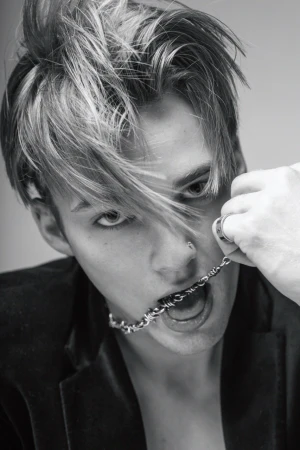 Black and white portrait of a young adult male with bleached hair holding a metal chain in his mouth wearing a blazer jacket.