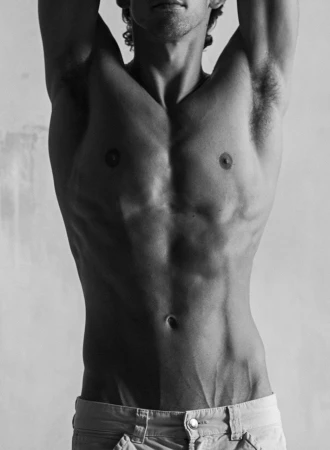 Black and white close-up studio shot of a muscular adult male torso with hands raised, revealing toned abdomen and partial face.