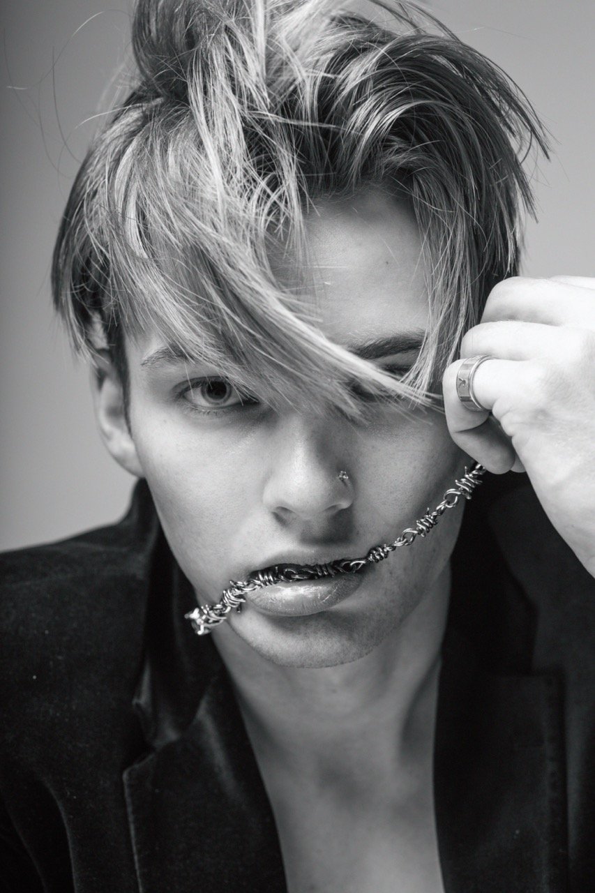 Black and white portrait of a young adult male with a barbed wire necklace, striking a fashionable pose indoors, wearing a velvet jacket.