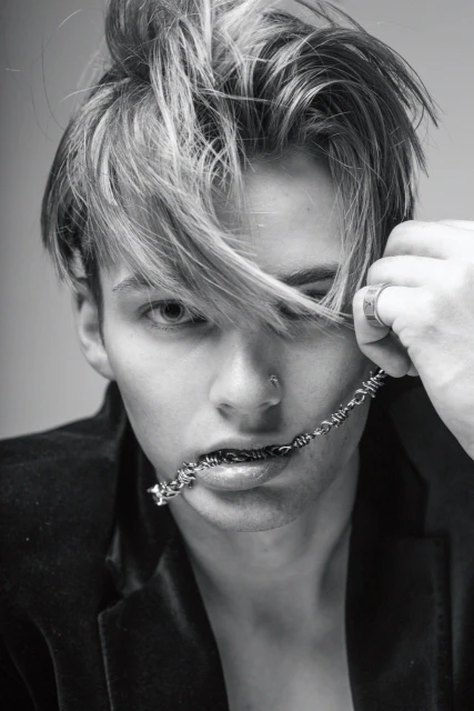 Black and white portrait of a young adult male with a barbed wire necklace, striking a fashionable pose indoors, wearing a velvet jacket.