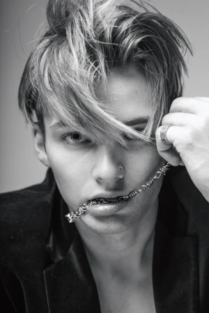 Black and white portrait of a young adult male with a barbed wire necklace, striking a fashionable pose indoors, wearing a velvet jacket.