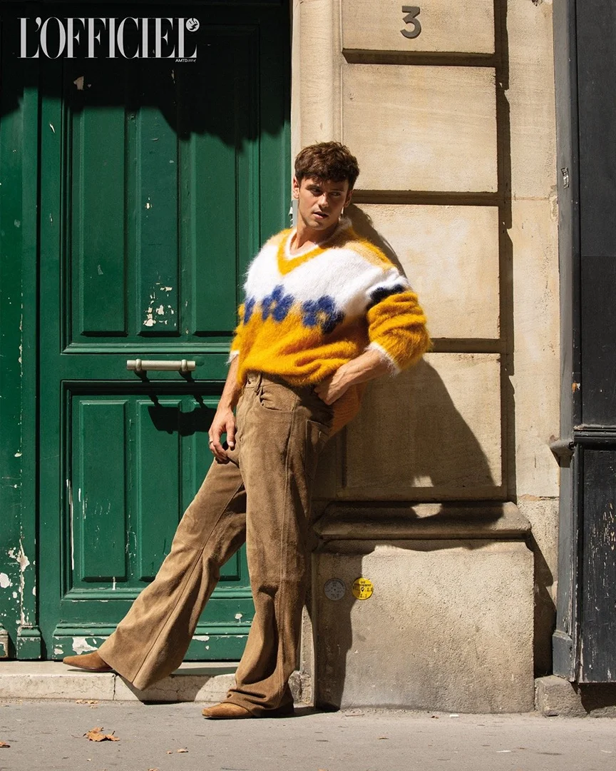 A casually dressed man leans against a stone wall next to a green door in natural light, featuring a sweater and tan corduroy pants.