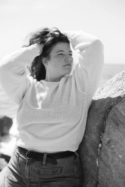 Black and white portrait of a woman with freckles wearing a sweater, leaning against rocks near the ocean on a sunny day.