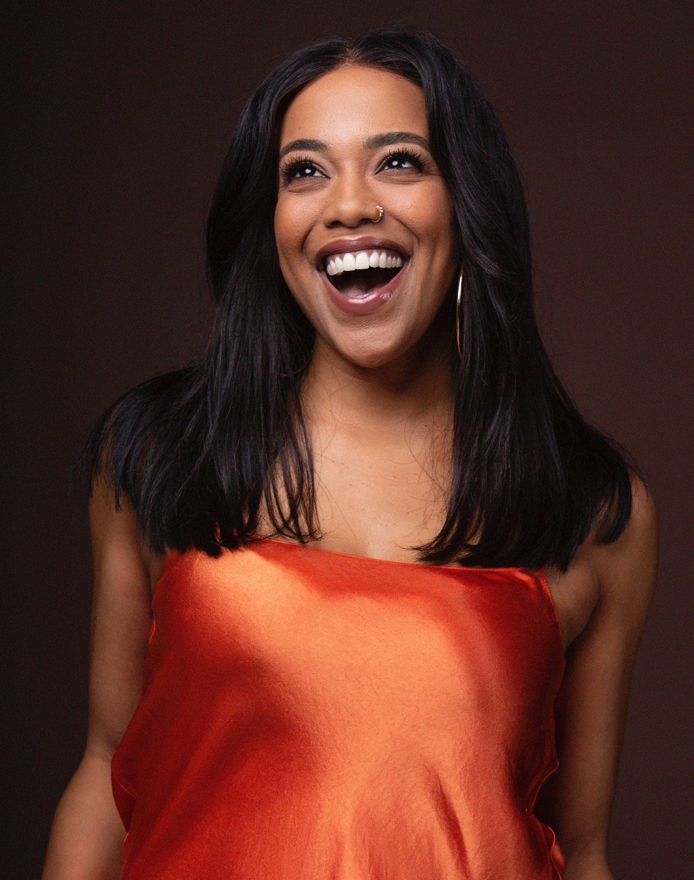 Portrait of a smiling young woman with dark hair wearing a bright orange top against a brown background, looking off-camera.