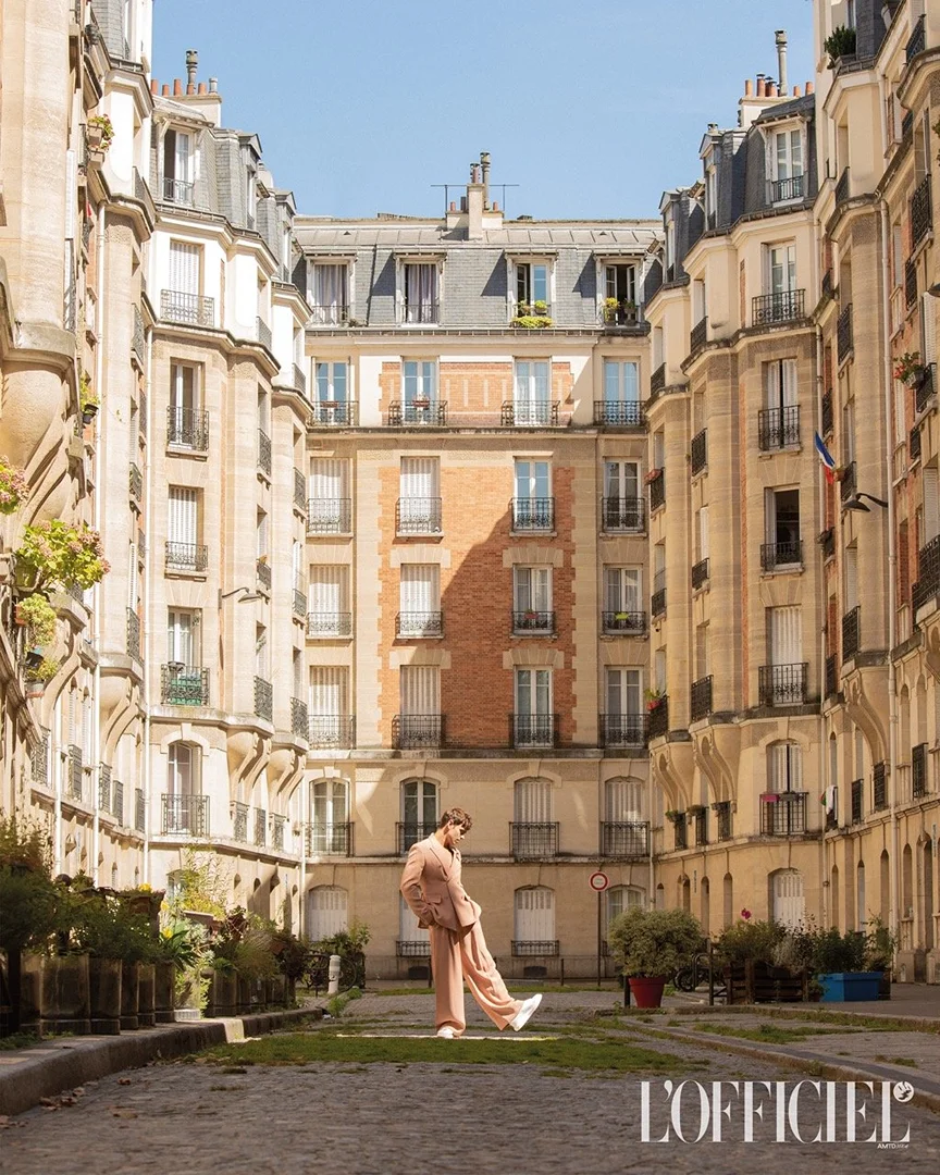 A stylish person in a light pink suit poses in a cobblestone courtyard surrounded by tall European buildings on a sunny day in an editorial fashion shoot.